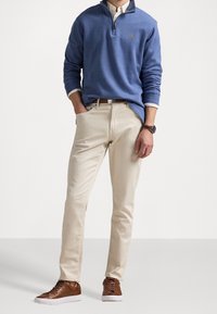 Sudadera de medio cierre en azul claro, chinos crema y zapatillas marrones. La sudadera presenta un pequeño logo, mientras que las zapatillas tienen una suela y cordones blancos.
