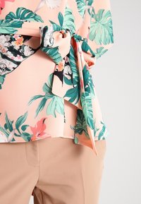 Blusa floreale in pesca con foglie tropicali verdi e fiori rosa, caratterizzata da un dettaglio in vita legato e un fit morbido. Abbinata a pantaloni beige.