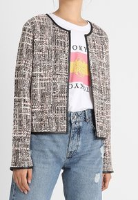 Marynarka w stylu tweed w odcieniach różu i szarości, z okrągłym dekoltem i czarnymi wykończeniami, w zestawieniu z koszulką graficzną i jasnoniebieskimi dżinsami.