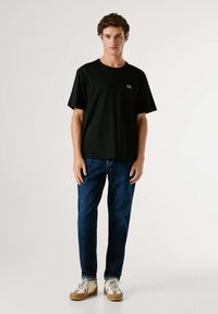 Camiseta negra de manga corta con el logo PJL, combinada con jeans de mezclilla azul y zapatillas color caramelo con detalles blancos y cordones texturizados.