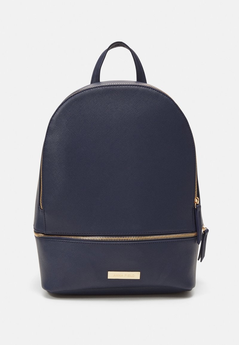 Sac à dos en cuir bleu foncé avec des fermetures éclair dorées, une poignée supérieure et une petite plaque rectangulaire dorée avec logo sur fond blanc uni.