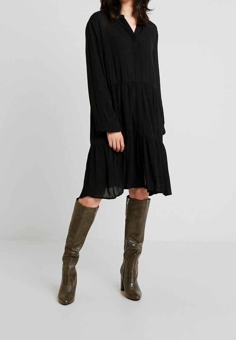 Femme portant une robe longue noire à volants à manches longues et des bottes à talons texturées vert olive montant jusqu'aux genoux, debout devant un fond clair uni.