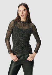 Donna che indossa una blusa trasparente a maniche lunghe con stampa animalier e pantaloni in pelle nera, con le mani sui fianchi contro uno sfondo semplice.