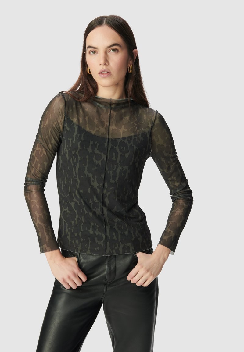 Femme portant un haut en imprimé animal à manches longues transparent et un pantalon en cuir noir, les mains reposant sur les hanches devant un fond uni.