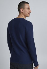 Temno modra dolgosleeve pulover s teksturiranim rebrastim vzorcem, okroglo ovratnikom in oprijetim krojem, nošen s strani.