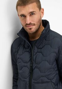 Giacca imbottita navy con colletto alto, dotata di zip frontale e motivo esagonale. Tessuto strutturato con tasche laterali.