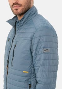 Hellblaue Steppjacke mit hohem Kragen, Reißverschluss vorne und horizontalen Nähten. Verfügt über eine Brusttasche mit orangefarbenem Akzent und Logo-Patch.