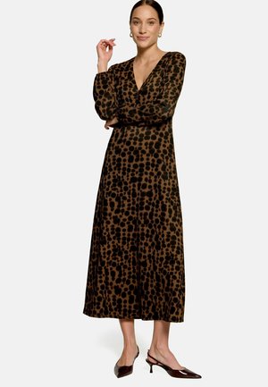 Femme portant une longue robe marron avec des taches noires irrégulières et des manches longues, debout, un bras croisé et l'autre main levée.