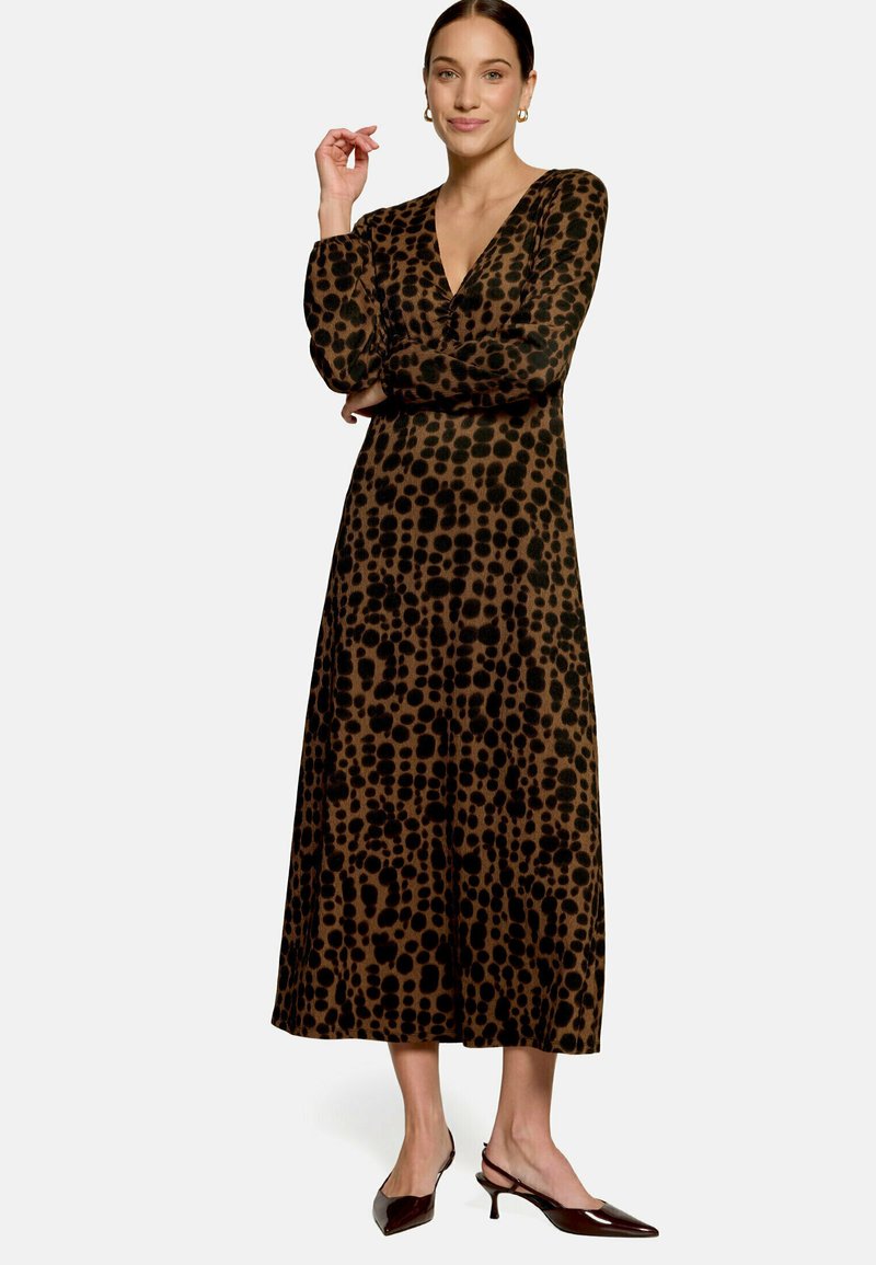 Femme portant une longue robe marron avec des taches noires irrégulières et des manches longues, debout, un bras croisé et l'autre main levée.