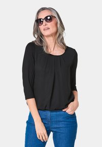Schwarze Bluse mit Dreiviertelärmeln, rundem Ausschnitt und Falten-Detail; kombiniert mit blauen Jeans und großen schwarzen Sonnenbrillen.