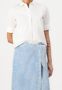 Femme portant une chemise blanche à manches courtes boutonnée, rentrée dans une jupe portefeuille en jean bleu clair avec un détail de nœud à l'avant.