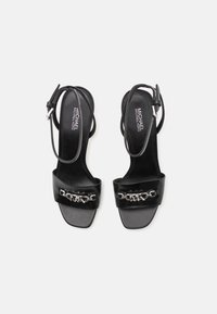 Black high-heeled sandals with a patent leather finish, featuring a wide strap adorned with a silver chain accent and thin ankle straps.