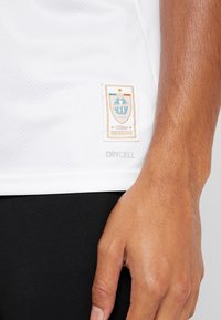 Valkoinen urheilu paita, jossa on teksturoitu kangas ja kudottu logopatchi, jossa lukee "120. vuosipäivä" sinisissä ja oransseissa yksityiskohdissa.