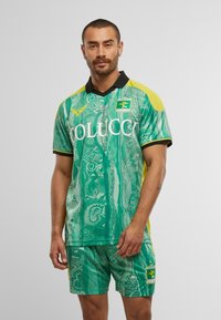 Mann in einem grünen und gelben Fußballtrikot mit den Logos "COLUCCI" und "Fussball Club Kitzbühel", der vor einem schlichten Hintergrund steht.