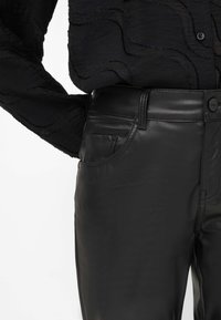 Pantalon en cuir synthétique noir avec une texture lisse, dotée d'une poche avant et d'une taille standard. Porté avec un haut à manches longues noir texturé.