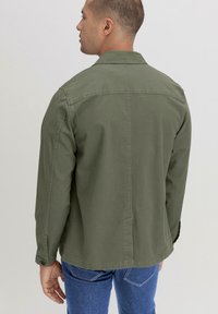 Olijfgroene, lichte katoenen jas met een rechte snit, met knoopsluiting bij de mouwen en een kraag. Zichtbare stiksels en minimalistische ontwerpelementen.