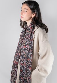 Sciarpa nera decorata con funghi di vari colori; tessuto leggero che si drappeggia in modo elegante. Il modello indossa un cappotto oversize di colore crema.