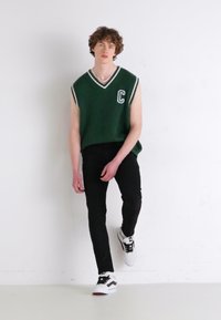 Zielony prążkowany sweater bez rękawów z białymi akcentami i literą "C", czarne obcisłe dżinsy i czarno-białe sneakersy. Gładkie tło.