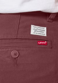 Gros plan de la poche d'un pantalon Levi's bordeaux montrant des étiquettes blanches et rouges de la marque Levi's et un bouton avec une partie d'une chemise blanche au-dessus.