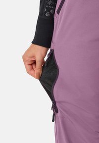 Hand die zwarte gaas ventilatiepaneel met dubbele ritsen aan de zijkant van paarse outdoorbroeken aanpast, terwijl een zwart shirt met sneeuwvlokpatroon wordt gedragen.