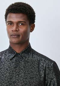 Czarna koszula w wzory z subtelnym nadrukiem zwierzęcym, z fasonem ze strefą sztywną i zapięciem na guziki. Teksturowany materiał ma połysk.