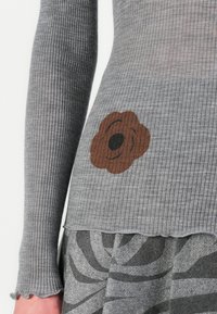 Grå ribbestrikket genser med bølget nederkant, med et brunt blomstermønster på siden. Stoffet ser mykt og teksturert ut.