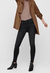 Jean skinny noir taille haute, associé à une veste en daim marron et un haut beige texturé. Bottines noires avec des accents argentés.