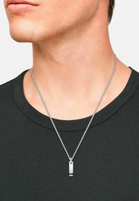 Close-up van een persoon die een zilveren ketting met een rechthoekige metalen hanger draagt boven een zwart T-shirt met ronde hals.