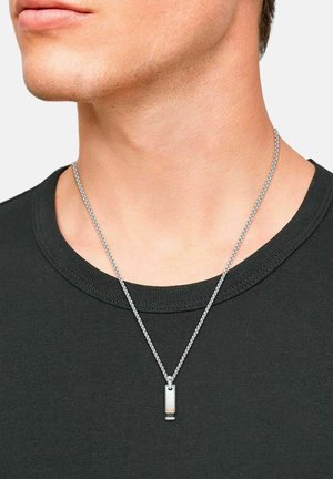 Nahaufnahme einer Person, die eine silberne Kettenhalskette mit einem rechteckigen metallischen Anhänger über einem schwarzen Rundhals-Shirt trägt.