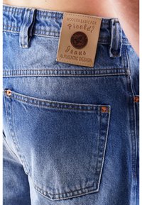 Niebieskie dżinsy z denimu z naszywką ze skóry z wytłoczonym tekstem. Widoczne przeszycia i miedziane elementy ozdobne na kieszeniach.