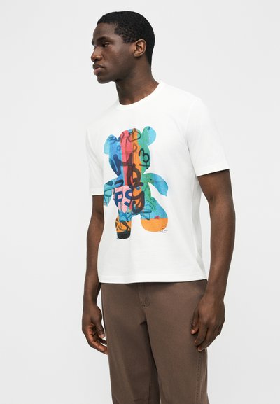 Άνδρας που φοράει λευκό μπλουζάκι με πολύχρωμο αφηρημένο σχέδιο αρκούδας και καφέ παντελόνι, στραμμένος αριστερά σε απλό γκρίζο φόντο.