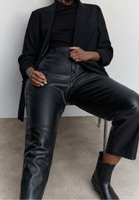 I pantaloni in pelle nera presentano una vita alta e una texture liscia, abbinati a un fitted dolcevita nero e a un blazer oversize. Stivaletti neri alla caviglia completano l'outfit.