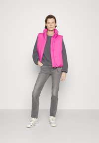 Gilet imbottito rosa brillante con colletto alto, maglione grigio strutturato, jeans grigi e sneakers bianche, in piedi su uno sfondo neutro.