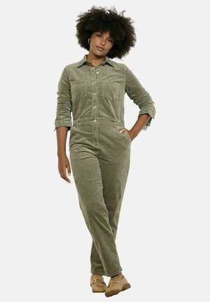Olijfgroene corduroy jumpsuit met lange mouwen, knoopsluiting, borstzakken en taps toelopende benen, gecombineerd met lichtbruine schoenen.