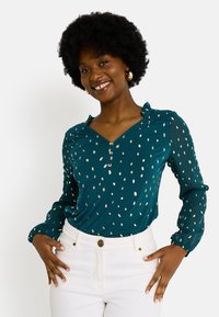 Blouse teal avec décolleté en V, manches longues et poignets froncés. Présente des motifs de pois dorés métalliques et une patte de boutonnage. Associée avec un pantalon blanc.