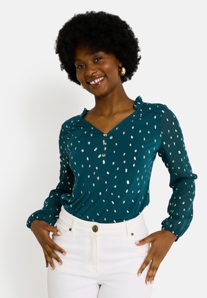 Blouse teal avec décolleté en V, manches longues et poignets froncés. Présente des motifs de pois dorés métalliques et une patte de boutonnage. Associée avec un pantalon blanc.