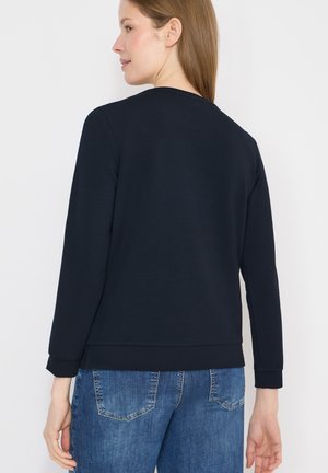 Femme vue de dos, portant un haut bleu marine à manches longues et un jean bleu, sur un fond blanc uni.