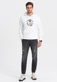 Sudadera blanca con capucha y gráfico en el frente, combinada con jeans negros desgastados y zapatillas deportivas gris claro, con puños elásticos y cordones.