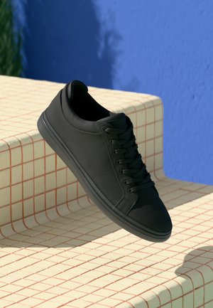 Black leather sneaker with a textured upper, flat laces, and a rounded toe. Features a subtle seam design and a textured rubber sole.
