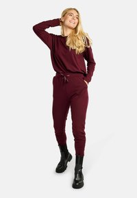 Vijoličasto rdeča majica z dolgimi rokavi in okroglim izrezom ter ujemajoče se športne hlače z elastiko v pasu, elastičnimi manšetami in črnimi gležnjarji.