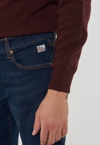 Maglione a maglia bordeaux con maniche lunghe abbinato a jeans in denim blu scuro. I jeans presentano un'etichetta bianca con testo rosso sulla tasca posteriore.