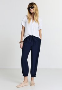 Chemise blanche à manches courtes avec boutons, pantalon bleu marine à cordon de serrage avec boutons-pression, et mocassins beiges. Les accessoires comprennent des bracelets en bois et des lunettes de soleil.