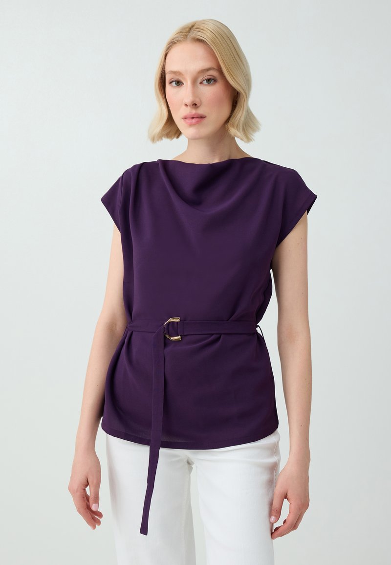 Femme au carré blond, portant un haut violet sans manches ceinturé et un pantalon blanc, debout devant un fond clair uni.