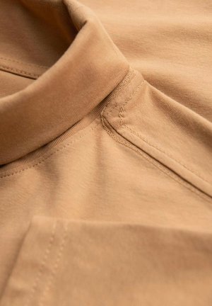 Tan bomullstyg med en slät yta, med en vikbar krage och synliga sömmar. En sidoficka är delvis synlig på vänster sida.