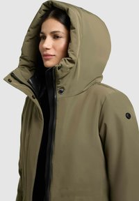 Chaqueta con capucha de color verde oliva y cuello alto, fabricada con un material impermeable, con cierres de cremallera y automáticos, y detalles en negro; diseñada para proporcionar calor y protección.