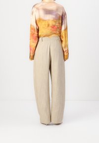 Femme portant un pantalon large beige et un haut à manches longues avec un motif flou multicolore, se tenant contre un fond blanc uni.