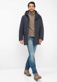 Man loopt en draagt een blauwe winterjas over een brown coltrui, lichte spijkerbroek en brown suède laarzen op een witte achtergrond.