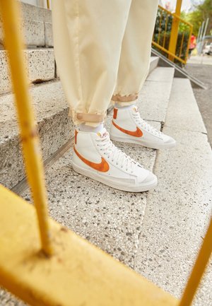Białe wysokie sneakersy z pomarańczowym logo Nike, noszone z beżowymi spodniami, stojące na zewnętrznych betonowych schodach obok żółtej poręczy.