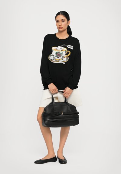 Schwarzer Pullover mit einer Teetasse-Grafik und floralen Details. Das Modell hält eine schwarze Lederhandtasche mit Trägern. Weiße Rock und schwarze Ballerinas.