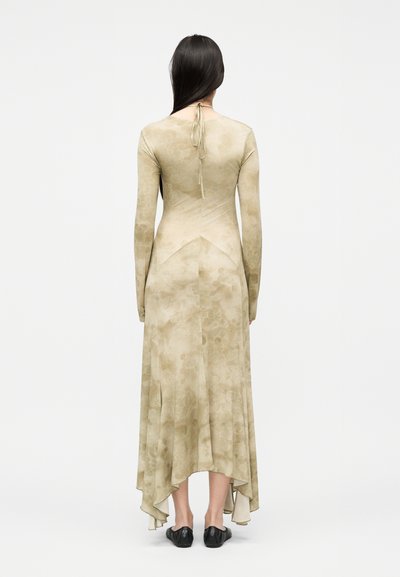 Γυναίκα με μακριά σκούρα μαλλιά που φοράει μπεζ φόρεμα με μακριά μανίκια, με φιόγκο στο λαιμό και ασύμμετρο τελείωμα, στέκεται με τα ποδιά της στραμμένα προς τα πίσω, φορώντας μαύρα παπούτσια.